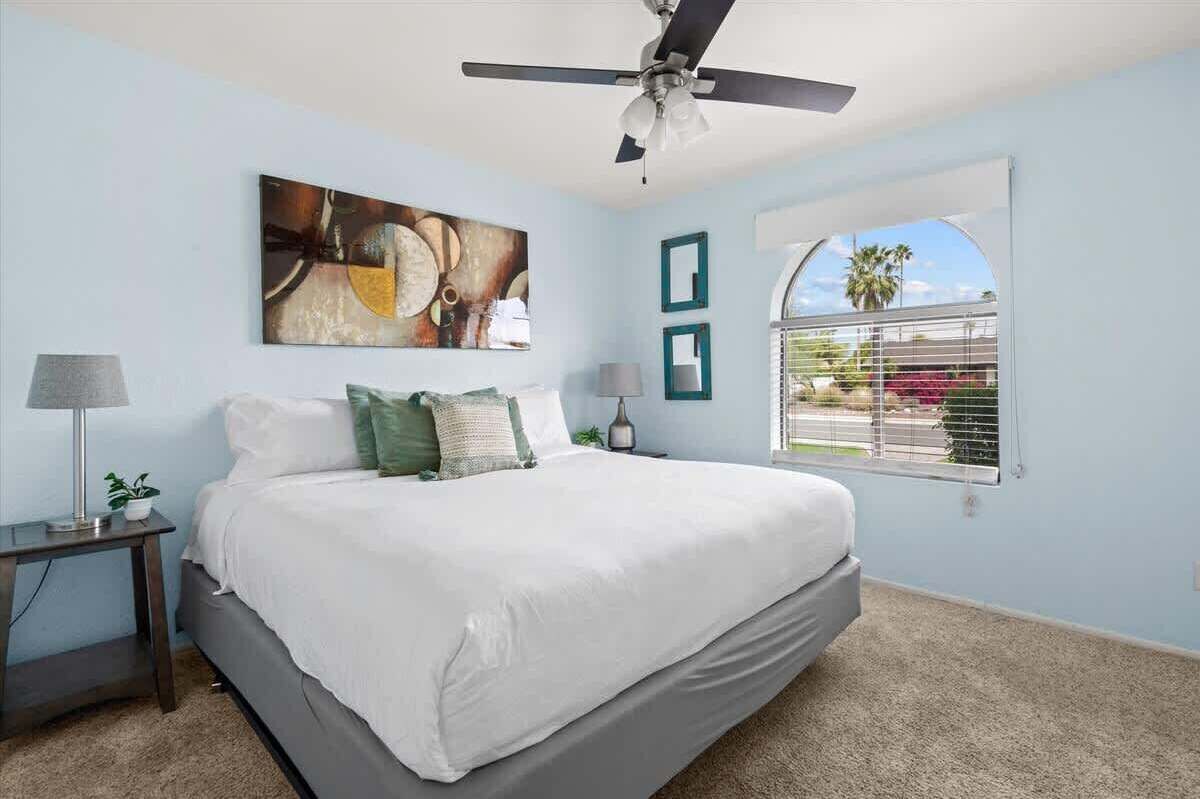A bedroom with a bed, a ceiling fan, a window with a view, and a painting on the wall.