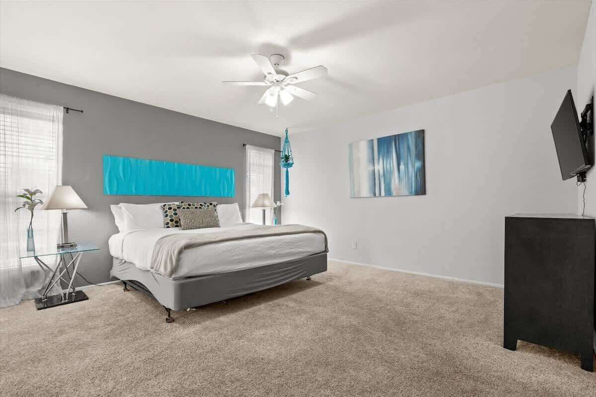 A modern bedroom with a bed, a ceiling fan, a TV, and a painting on the wall.