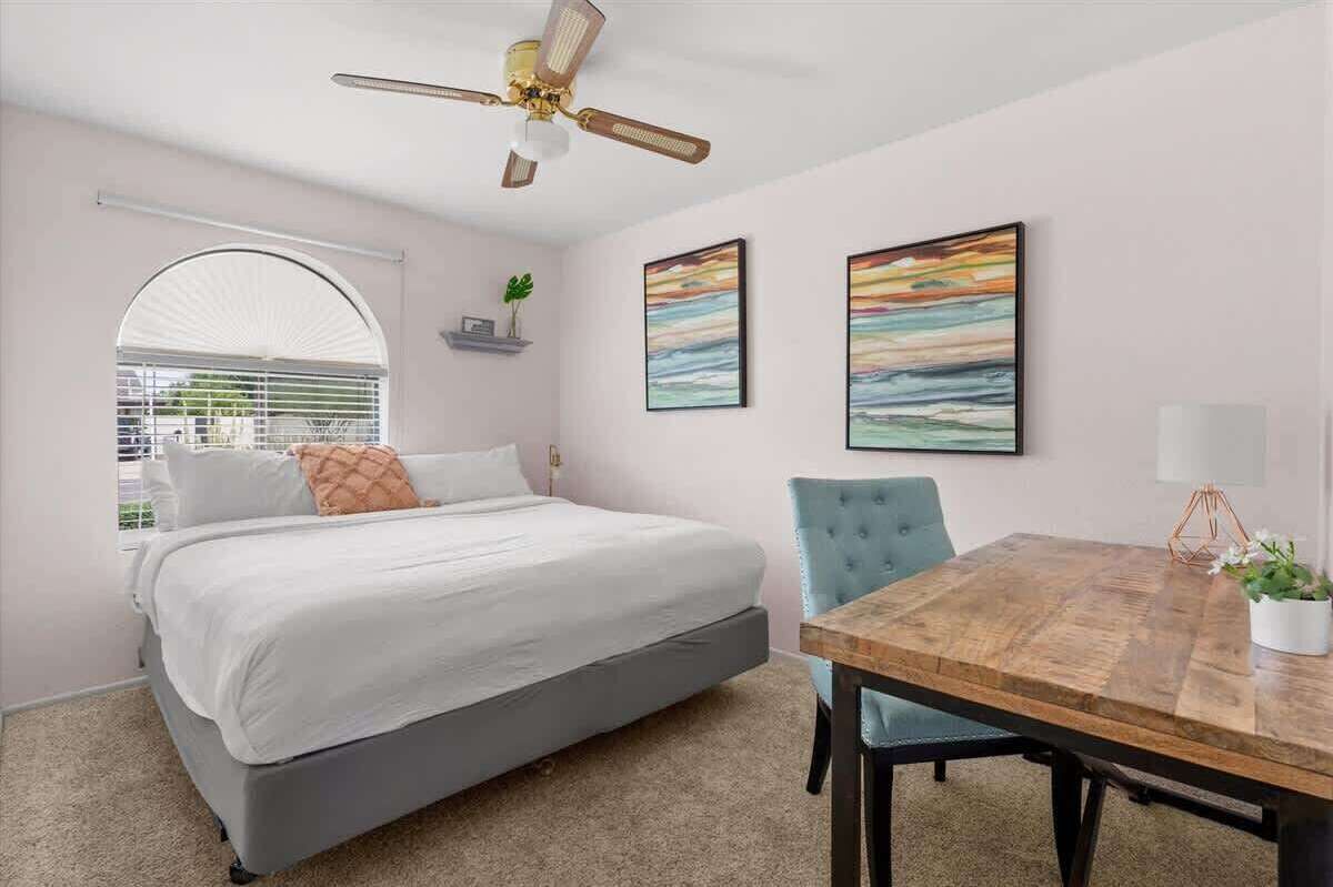 Un dormitorio con cama, ventilador de techo, mesa de comedor con sillas y dos cuadros en la pared.