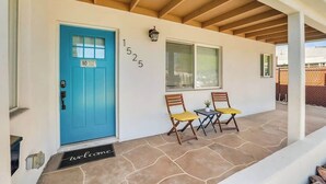Eine blaue Tür mit einem Schild 'NO Soliciting', Hausnummer 1525, und zwei hölzerne Stühle mit gelben Kissen auf einer gepflasterten Veranda.