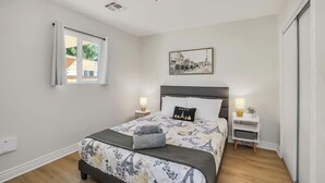 Een slaapkamer met een bed, nachtkastje, raam en een foto aan de muur.
