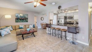 A modern living room with a sofa, coffee table, and a cityscape painting. A kitchen with bar stools and a ceiling fan is visible in the background. - Exclusive Tempe home: pool, BBQ, sleeps 8 (Tempe)