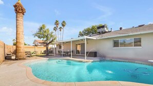 A swimming pool with a curved edge, a covered patio area with chairs, and a tall palm tree. - Exclusive Tempe home: pool, BBQ, sleeps 8 (Tempe)