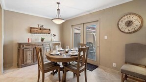 A dining area with a round table set for four, wooden chairs, a pendant light, a large wall clock, and a view through a glass door. - Exclusive 3BR Retreat, Pool & Spa Access (Scottsdale)