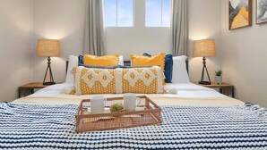Een net opgemaakt bed met een blauw-witte spreideken, twee bedlampjes en een dienblad met kopjes en een plantje.