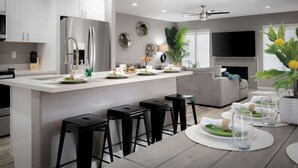 Een moderne keuken met een centraal eiland, zwarte barkrukken, een eettafel voor vier personen, en een zithoek met een bank en televisie.