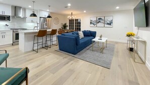 A modern living room with a blue sofa, a coffee table, and a TV. There is a kitchen area with white cabinets and a dining table. - Marbella Lane - Contemporary Sophisticated Home (Sunnyvale)