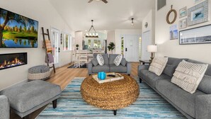 Een moderne woonkamer met een haard, grijze banken, een ronde geweven salontafel en decoratie met een nautisch thema.