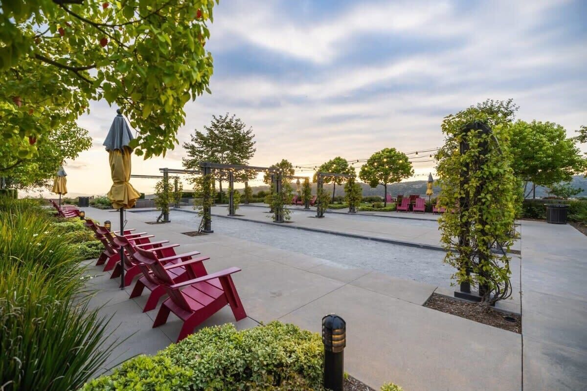 A spacious outdoor area with red benches, a pergola with string lights, and a variety of trees and shrubs.