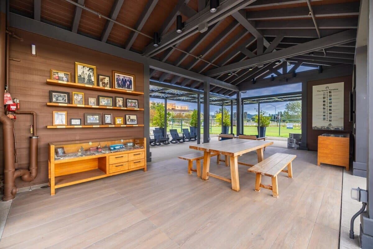 Un vaste espace extérieur aménagé avec un banc en bois, une grande table et des chaises. Des tableaux encadrés ornent le mur, et un extincteur est visible.