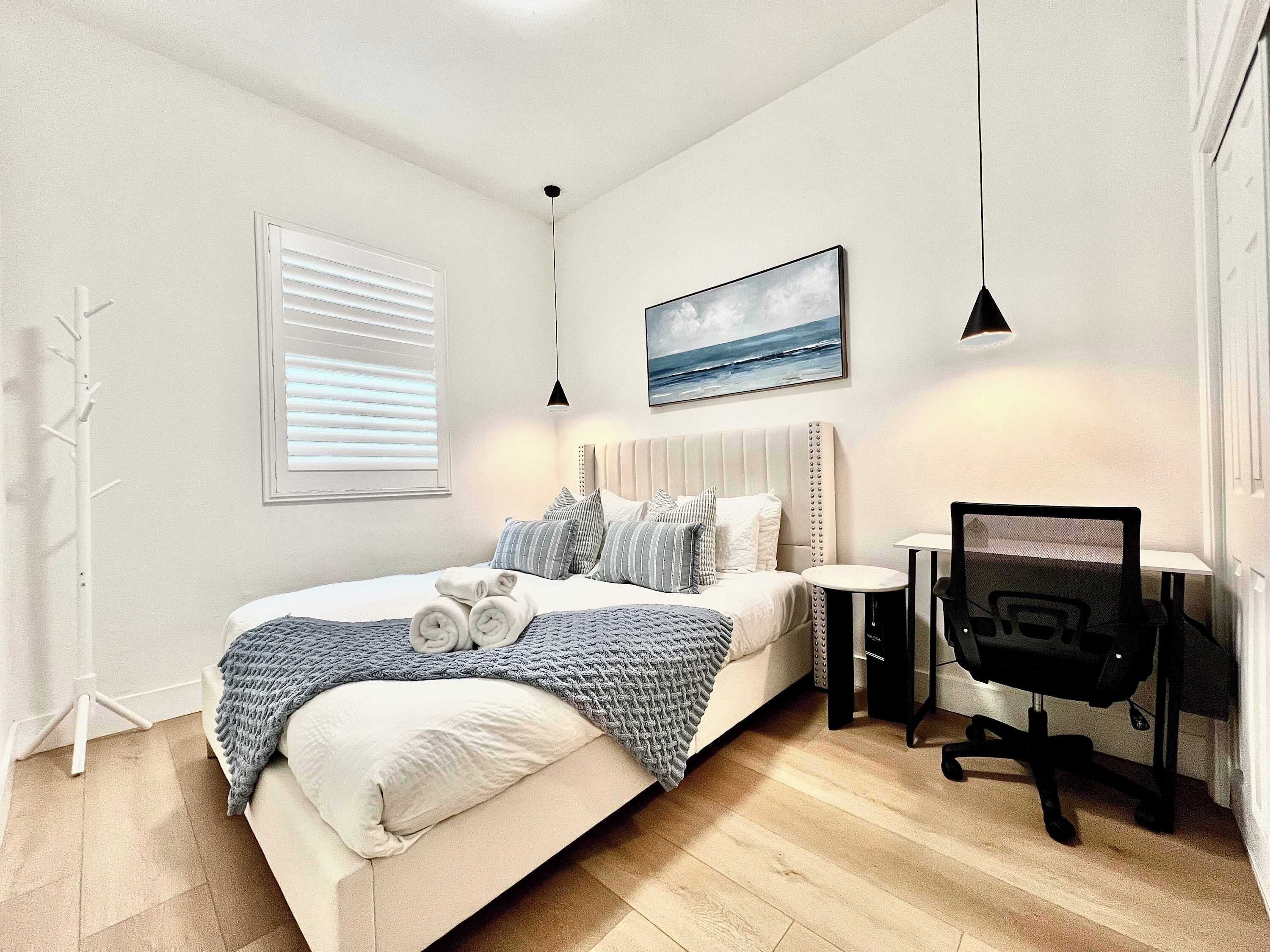 A bedroom with a bed, bedside table, desk, chair, and a painting on the wall.