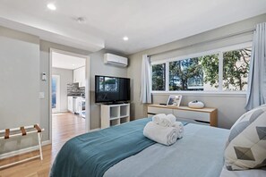 A modern hotel room with a large bed, a television, a window with a view of trees, and a small desk with a lamp. - Marbella Lane - Chic Abode w Mountain Skyline (Pacifica)
