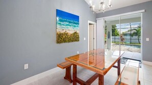 Zona pranzo con tavolo in vetro, un dipinto raffigurante una spiaggia e vista piscina attraverso porte in vetro.