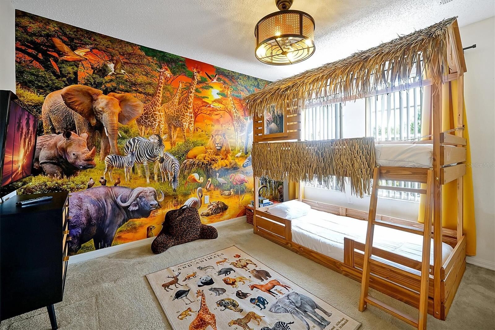A room with a mural of African wildlife, a bunk bed, a desk with a computer, and a window with a view of the outdoors.