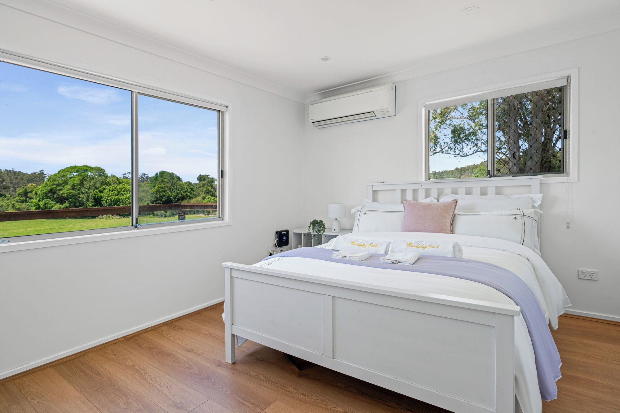 A bedroom with a large window, a bed with pillows, a bedside table, and an air conditioning unit.