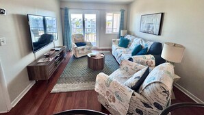 Un salon comprenant un canapé, un fauteuil, un téléviseur et donnant sur le balcon extérieur.