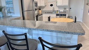 Une cuisine avec des meubles blancs, un plan de travail en granit, un réfrigérateur en acier inoxydable et une table à manger en bois.