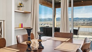 A dining area with a table set for four, a vase of flowers on a shelf, and a view of a distant landscape through large windows.