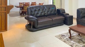 A spacious living room with a leather sofa, a dining area with wooden chairs, and a modern ceiling light fixture. - KIGALI QUEENS HOME AND APRTMENTS (Kigali)