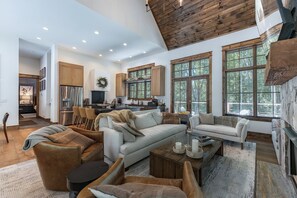 A spacious living area with a high ceiling, a fireplace, and large windows offering a view of the outdoors. - Luxe Eagles Nest Home: Hot Tub, 2 Outdoor Fireplaces & Fire Pit; Views (Banner Elk)