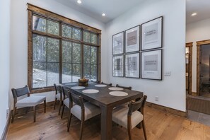 Uma área de refeições com uma mesa grande, seis cadeiras e vista para uma floresta através de amplas janelas.