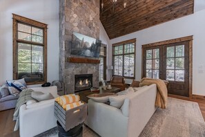 A cozy living room with a stone fireplace, large windows, and a high wooden ceiling. - Luxe Eagles Nest Home: Hot Tub, 2 Outdoor Fireplaces & Fire Pit; Views (Banner Elk)