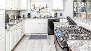 A modern kitchen with white cabinets, a gas stove, a dishwasher, and a window with a view of trees. - Spacious Home • Hot Tub • Sleeps 24 • Near Airport (Anchorage)