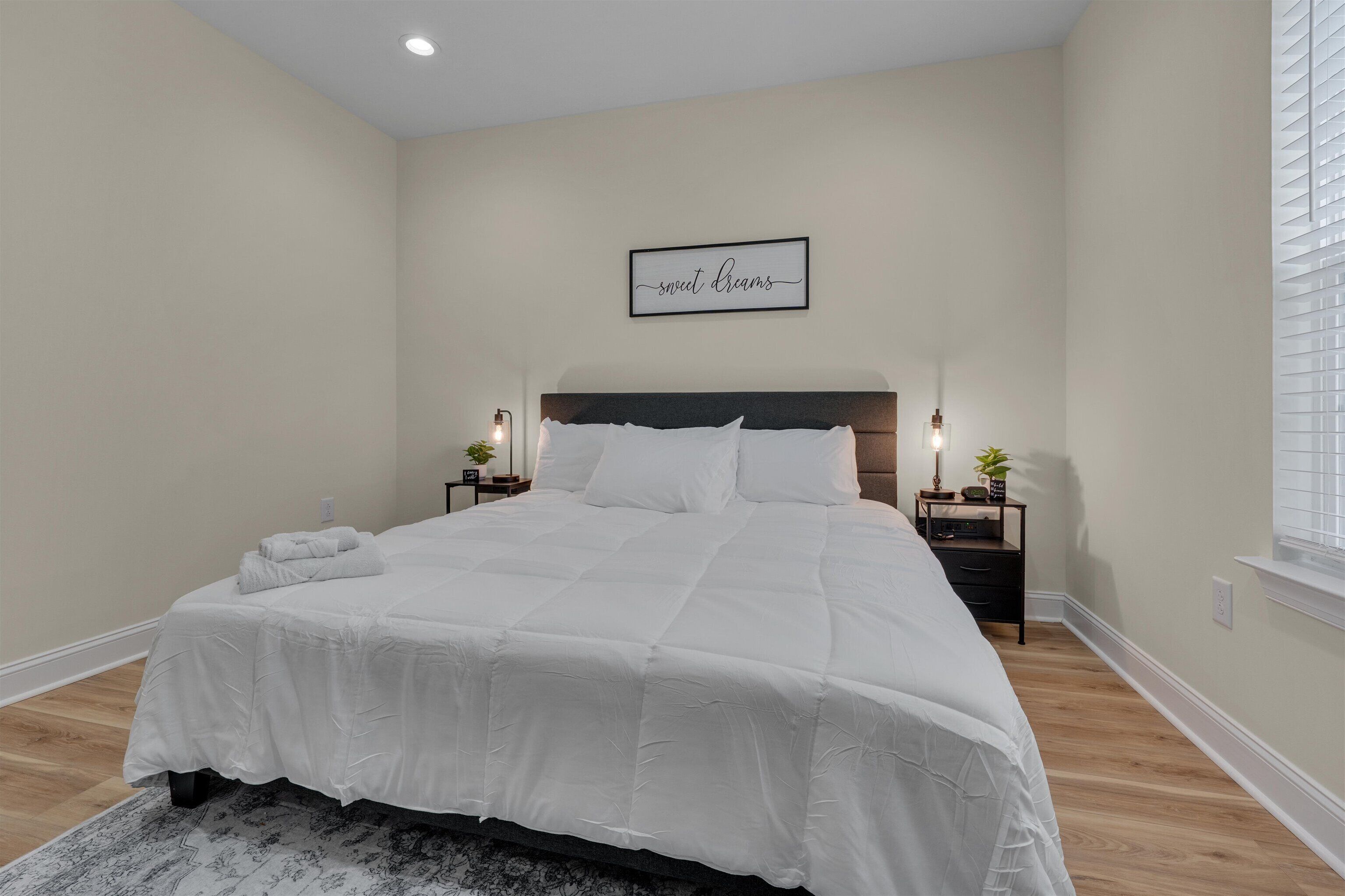 Une chambre à coucher avec un grand lit, deux tables de chevet avec des lampes, un panneau décoratif fixé au mur et une fenêtre avec des stores.