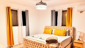 Ein Schlafzimmer mit einer gesteppten Kopfteile, einem Bett mit gelb-weißer Bettwäsche, einer Nachttischlampe und einem Holznachttisch.