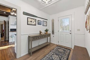 A hallway with a white door, a console table with decorative items, framed pictures on the wall, and a patterned rug. - Boat Builders House - Oceanfront Acadia Getaway (Tremont)