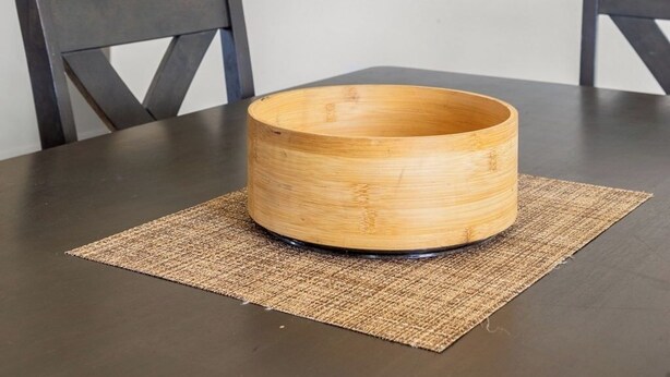Eine runde Holzkugel auf einem dunklen Tisch mit Untersetzer aus Bändern, zwei dunkle Stühle und eine schlichte weiße Wand.