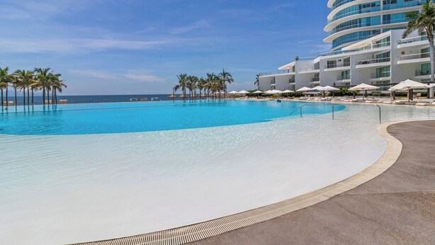Una gran piscina con borde curvo, rodeada de palmeras y un moderno edificio de hotel con múltiples balcones.