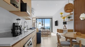 Una cocina moderna con horno empotrado y cafetera, un comedor con mesa y sillas de madera, y un ventanal con vistas al mar.
