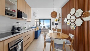 Cocina moderna con armarios de madera, horno empotrado y cafetera. Hay una zona de comedor con una mesa redonda y sillas, y vistas al océano a través de amplios ventanales.