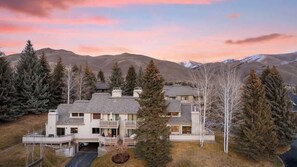 A large, multi-story house with a prominent balcony, surrounded by trees and mountains in the background.