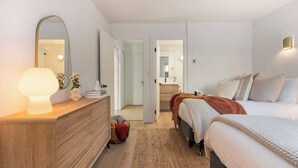 A bedroom with a bed, bedside table, lamp, dresser, mirror, and a door leading to another room.