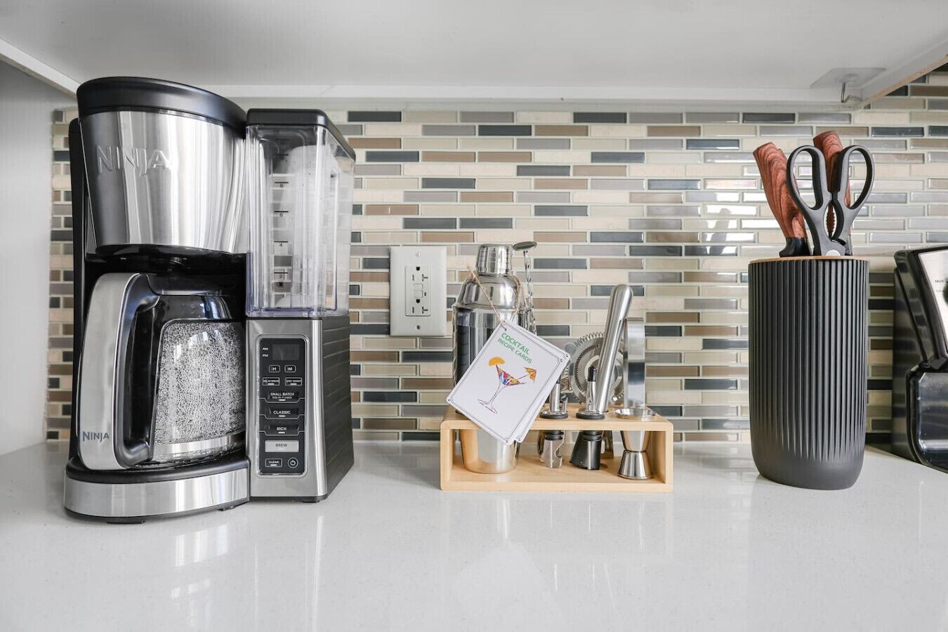 Une cuisine équipée d’une machine à café, d’un bloc à couteaux et d’une fiche contenant des recettes de cocktails, le tout posé sur le plan de travail.