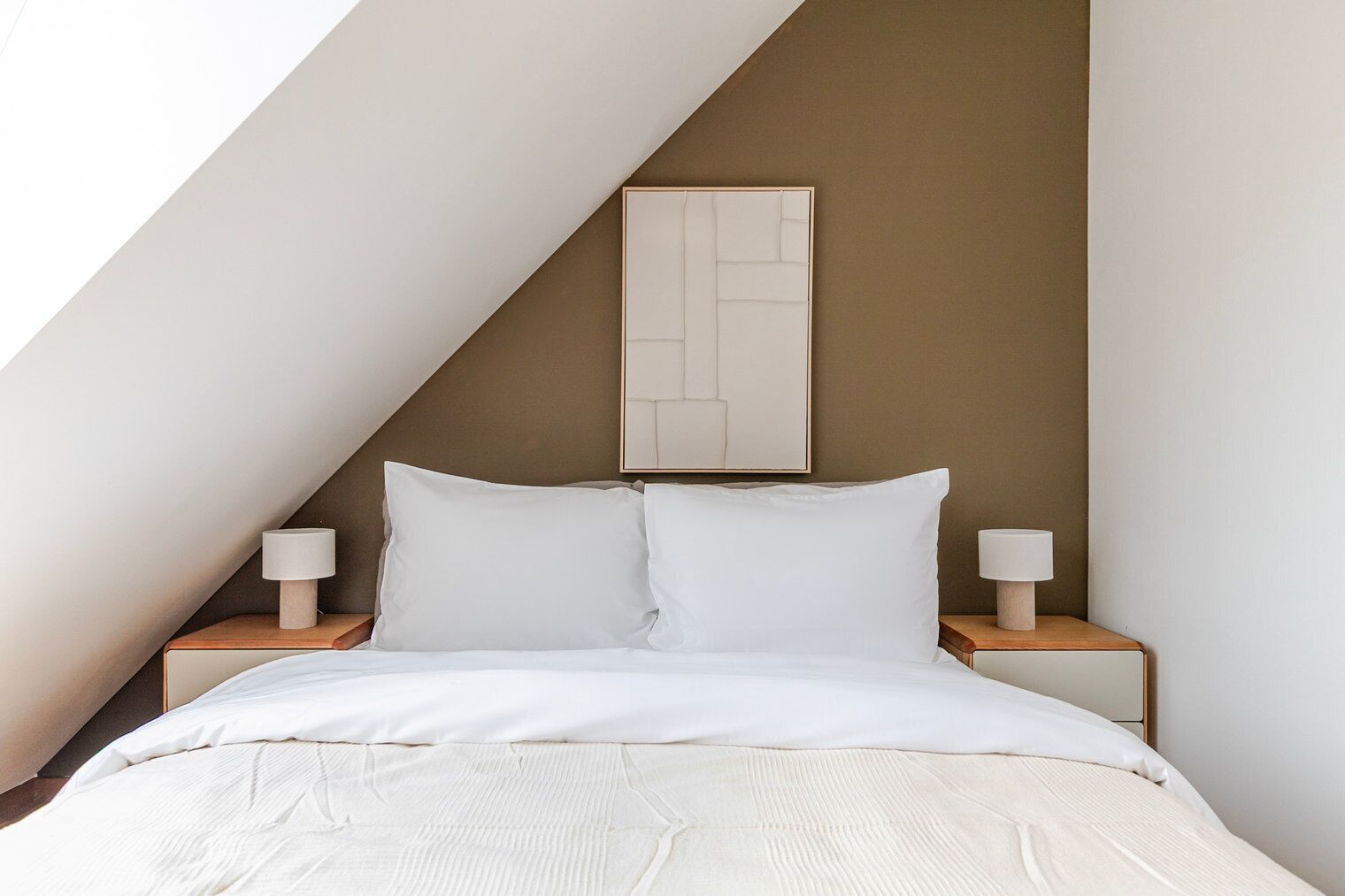 Ein Schlafzimmer mit schrägem Dach, einem Bett mit weißer Bettwäsche, zwei Nachttischen mit Lampen und einem gerahmten abstrakten Kunstwerk an der Wand.