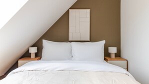 Une chambre avec un plafond en pente, un lit recouvert d’une literie blanche, deux tables de chevet avec des lampes et une œuvre d’art abstraite encadrée accrochée au mur.