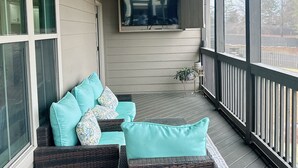 Eine überdachte Veranda mit Deckenventilator, einem an der Wand befestigten Fernseher und einer Sitzecke mit einem Rattan-Sofa und blaustichigem Sitzkissen.