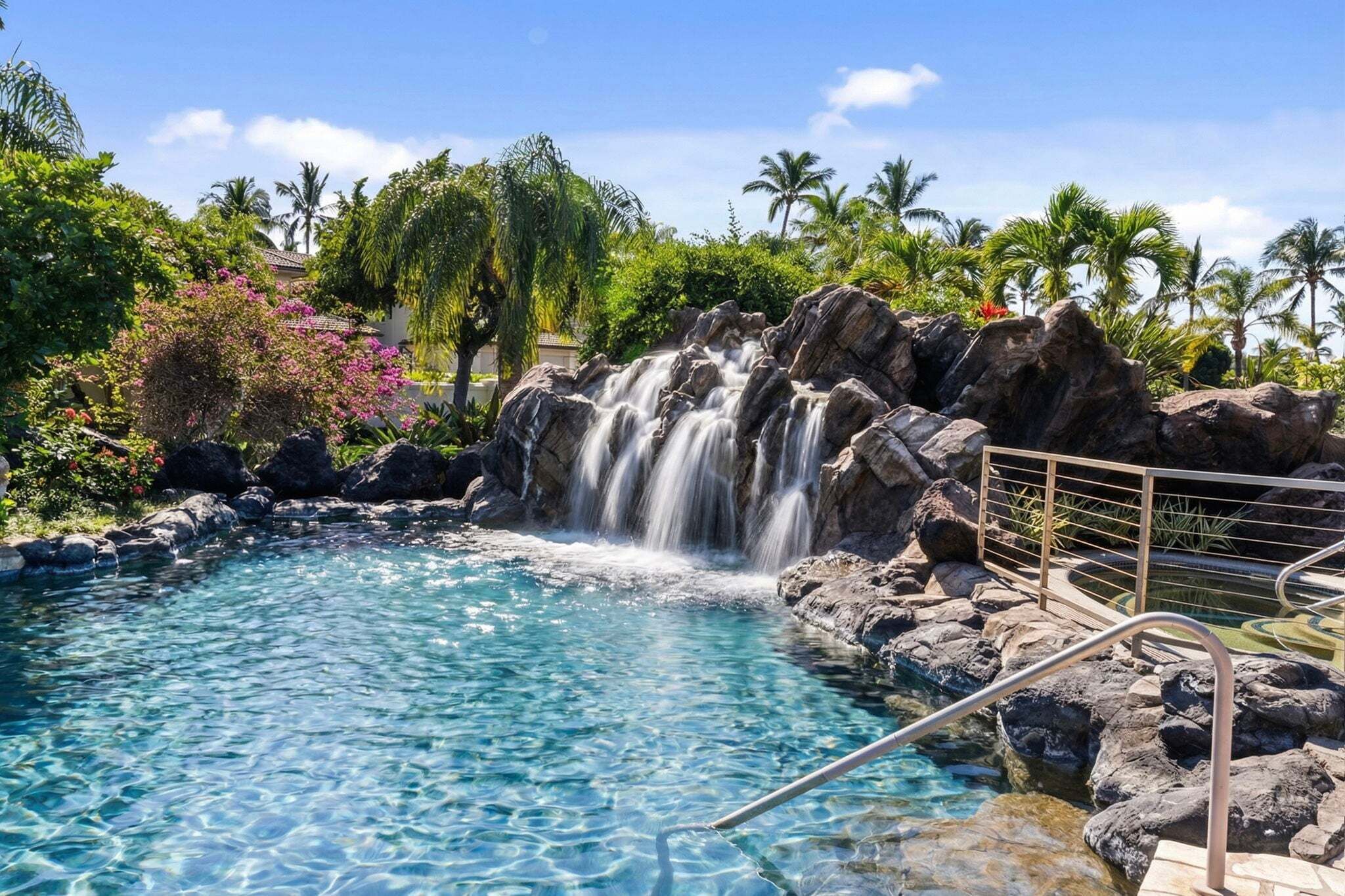 Een helder zwembad met een waterval, omringd door rotsen en palmbomen.