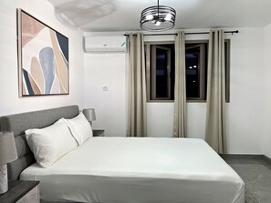 A hotel room with a bed, a nightstand, a lamp, a wall-mounted air conditioner, a window with curtains, and a ceiling light fixture. - Charming 1 Bedroom Apartment Unit in the Heart of The City. (Kingston)