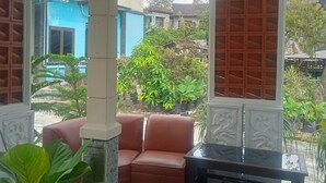 A covered outdoor seating area with a sofa and a table, surrounded by greenery and a view of a blue building in the background. - Pakemsari Resort Hotel (Pakem)