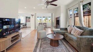 Ein geräumiges Wohnzimmer mit Flachbildfernseher, Deckenventilator, Sofa und Essbereich mit Tageslicht.