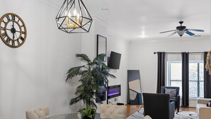 Una sala de estar moderna con una mesa de vidrio, asientos cómodos, una planta grande en maceta, un ventilador de techo y un televisor en la pared.