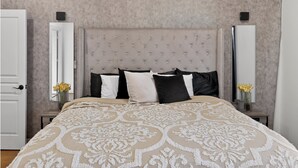 Un dormitorio con cabecera capitonada, una cama grande con colcha estampada, dos mesitas de noche con jarrones de flores y una puerta a la izquierda.