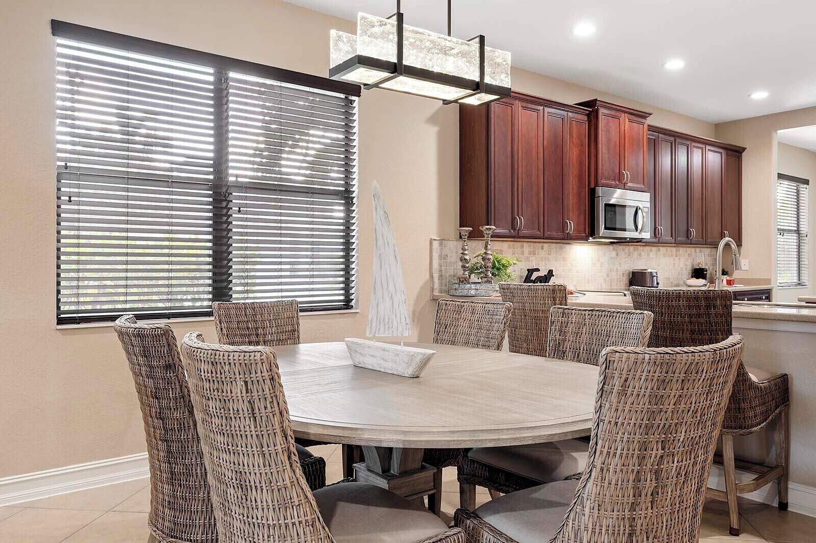 A dining area with a round table and wicker chairs, a kitchen with dark wooden cabinets, and a large window with blinds.