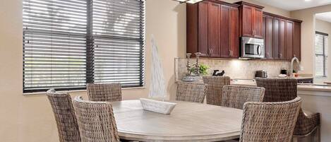 A dining area with a round table and wicker chairs, a kitchen with dark wooden cabinets, and a large window with blinds.