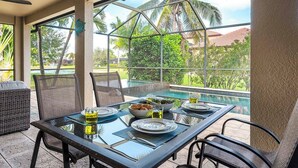 Une table à manger avec un plateau en verre, entourée de chaises, un panier de pain et un verre rempli d’une boisson, le tout mis en valeur par un décor composé d’une piscine et de palmiers.