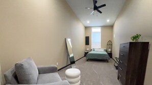 Quarto com sofá cinzento, um banquinho branco, cama com colcha verde, cômoda, espelho e ventilador de teto.